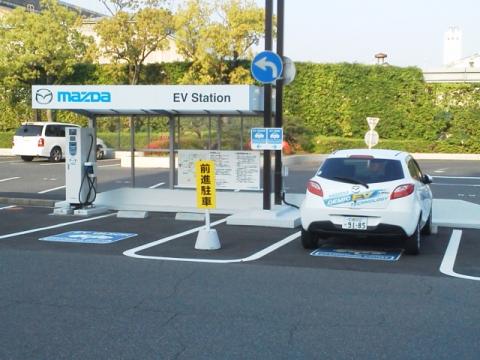 ここは，マツダの来客用駐車場に設けられたEVステーション，電気自動車の充電ステーションです．停まっているのはデミオの電気自動車です．この車の製造コストは・・・